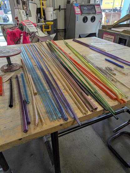 Blue, purple, brown, green, orange striped tubes laid out on a wooden table. Hot pink lathe and sandblaster in the background