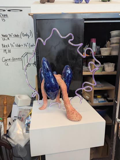 A ceramic, glass and Argon sculpture by the third year grad, Morgan. A ghostly purple whisp of glass encircles a dark blue glazed ceramic piece held up by a dripping terracotta mixed with clear glass oozing tripod on a white pedestal