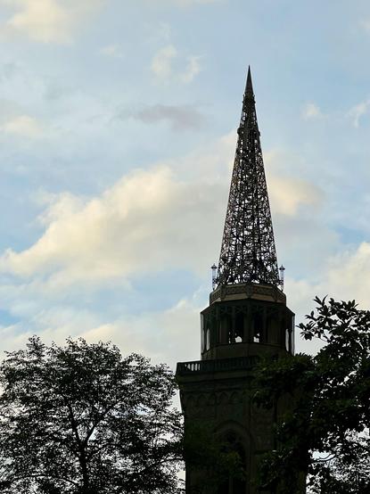 Spire at top of Smithfield United Church of Christ in Pittsburgh which is a latticework of metal. There appears to be a walking area below that and maybe a bell tower inside? The sky behind it (and seen through the lattice) is a blue sky with thin, fluffy clouds vying for a spot. If you were someone who saw faces in things then that cloud on the left surely looks like an aardvark.