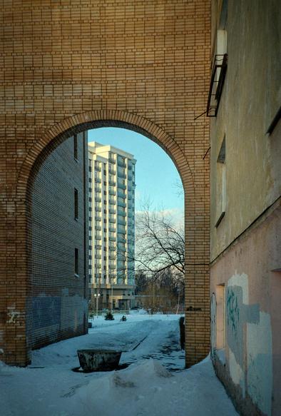 An archway of brick between the houses. In the arch one can see a multi-story house illuminated by the setting sun. On the right, in the foreground is a shabby wall of a house.