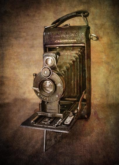 A vintage folding camera with intricate details and an accordion-style bellows is shown against a textured background, evoking a nostalgic and antique feel. The camera's lens and body showcase classic design elements and reflect early photographic technology.