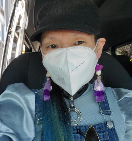 Selfie of me with blue-tipped hair wearing a white KN95 mask, a black newsboy cap, and purple tassel earrings with small skulls. They are sitting in what appears to be the passenger seat of a bus. They wear a leather choker with metal rings, a blue ribbed top, and denim overalls. One side of the scene includes a building outside the bus window.