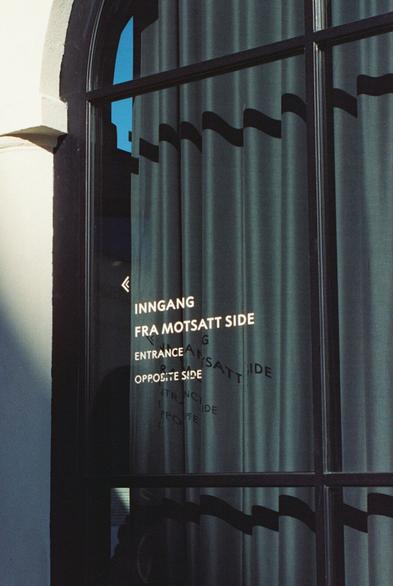 a window from the outside of a museum in oslo. there is text on the window that says "entrance opposite side" in norwegian and english. the frame of the window and the text cast shadows on the curtain behind them, making curvy lines and text on the curtain. it was a bright summer day when the photo was taken.