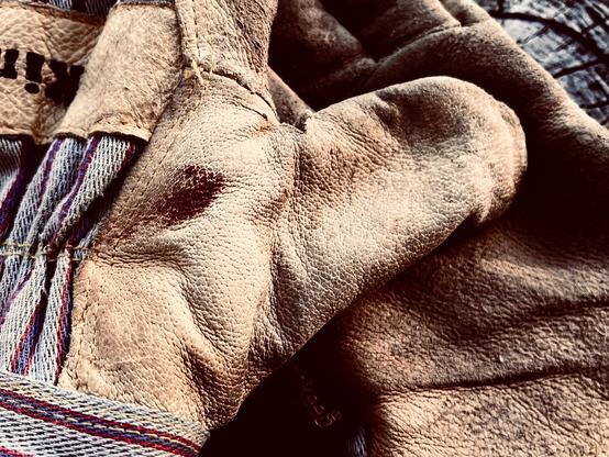 A close-up of a pair of well-worn leather work gloves. There is a smudge of blood on one glove near the thumb. The gloves seem to be resting on a chunk of dried wood as the end grain is visible.