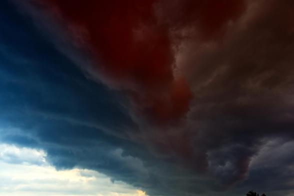 Ein Wolkenbild mit angehobenen Kontrasten. Eine keilförmige Wolkenformation schiebt sich von oben nach unten ins Bild. Links ist noch hellerer Himmel zu sehen. In der Mitte des Keils sind die Wolken am dicksten und hängen am tiefsten. Die untergehende Sonne färbt sie dunkelrot.