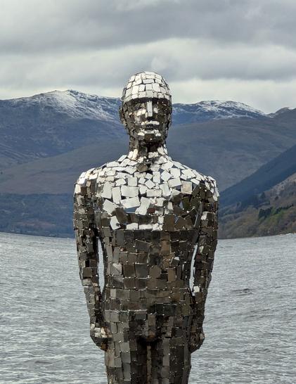 Image shows the metallic mirror man of Loch Earn in Scotland. 
Looking life sized near the shore. 
There's even some snow on the hills behind him too
